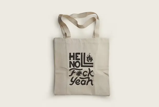 A canvas tote bag is laid out on a white surface, featuring the words “Hell No F*ck Yeah” in black on the front.